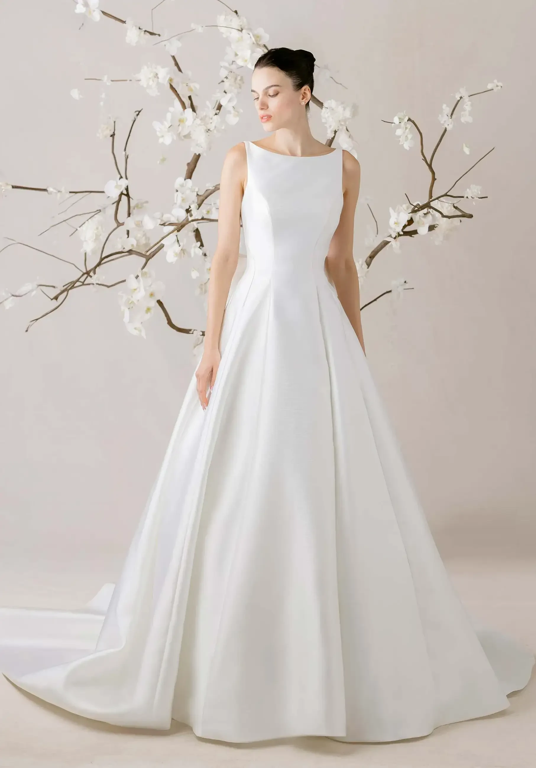 A woman in an elegant, sleeveless white gown stands gracefully in front of blooming branch decor. The scene conveys a serene, bridal elegance.