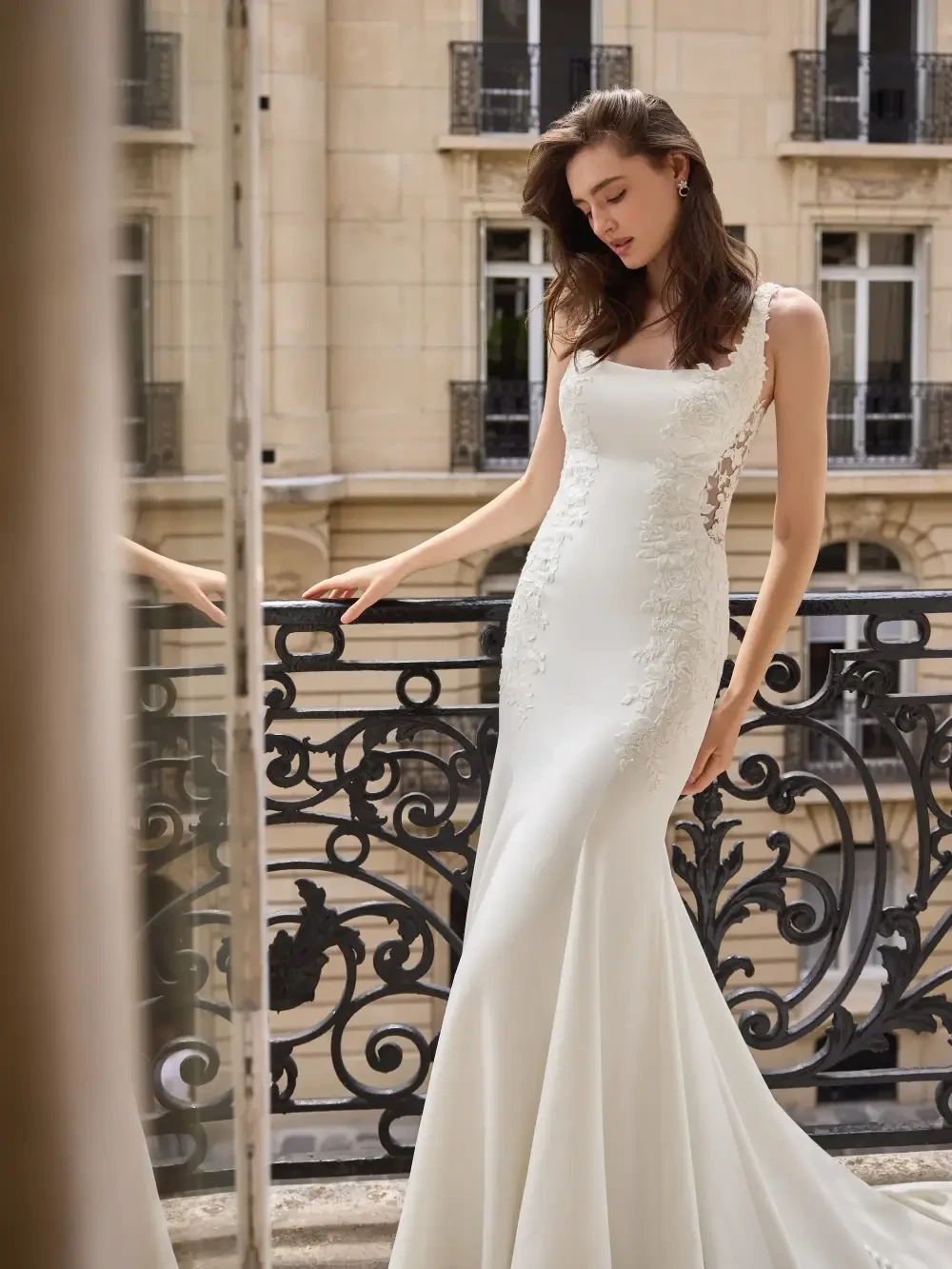 A woman in a white wedding dress stands elegantly near a decorative balcony railing, gazing down thoughtfully.