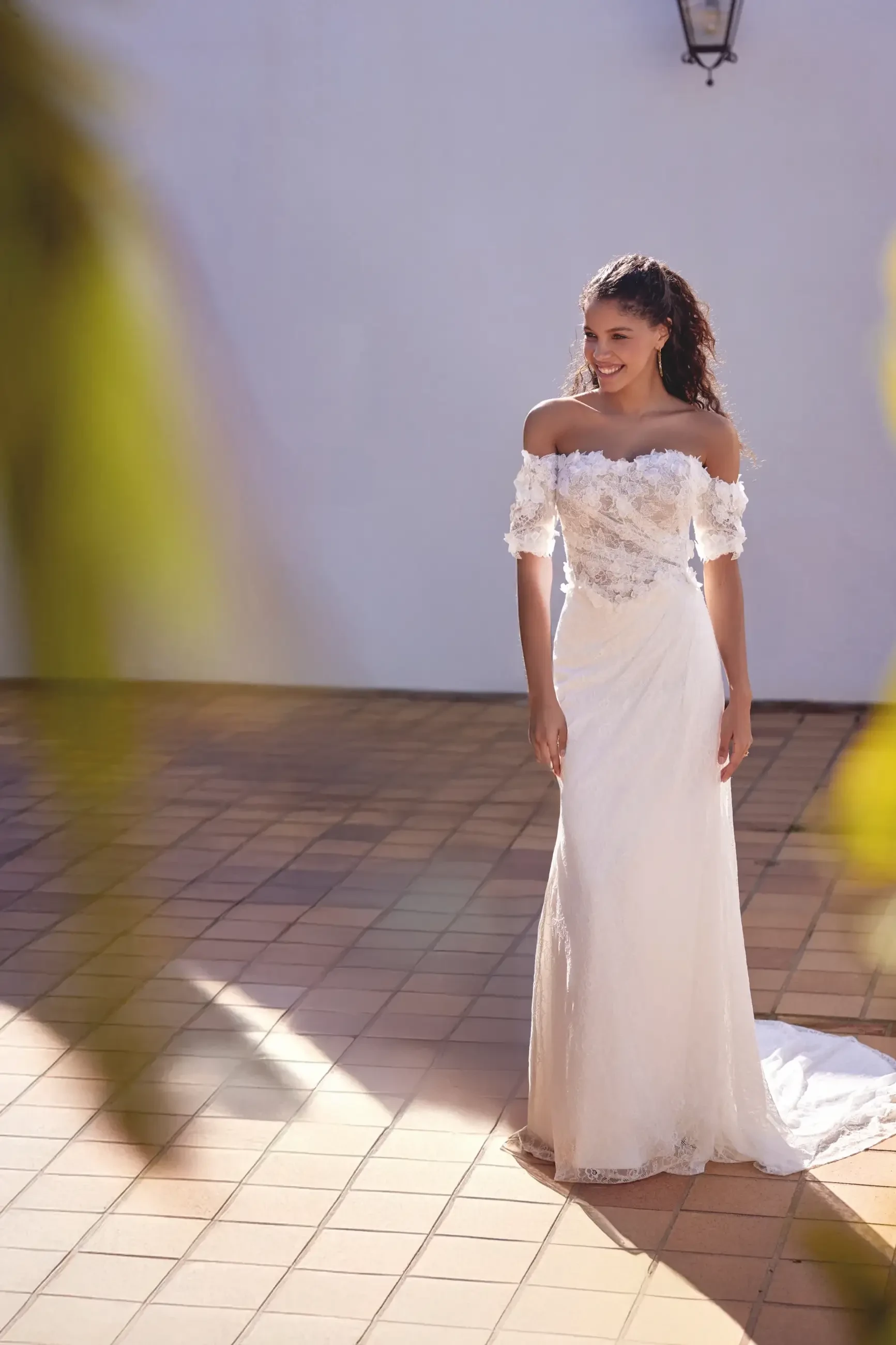 A woman in a bridal gown stands on a tiled surface with sunlight casting soft shadows. She has her hair down and smiles warmly, wearing an off-shoulder dress adorned with floral details.