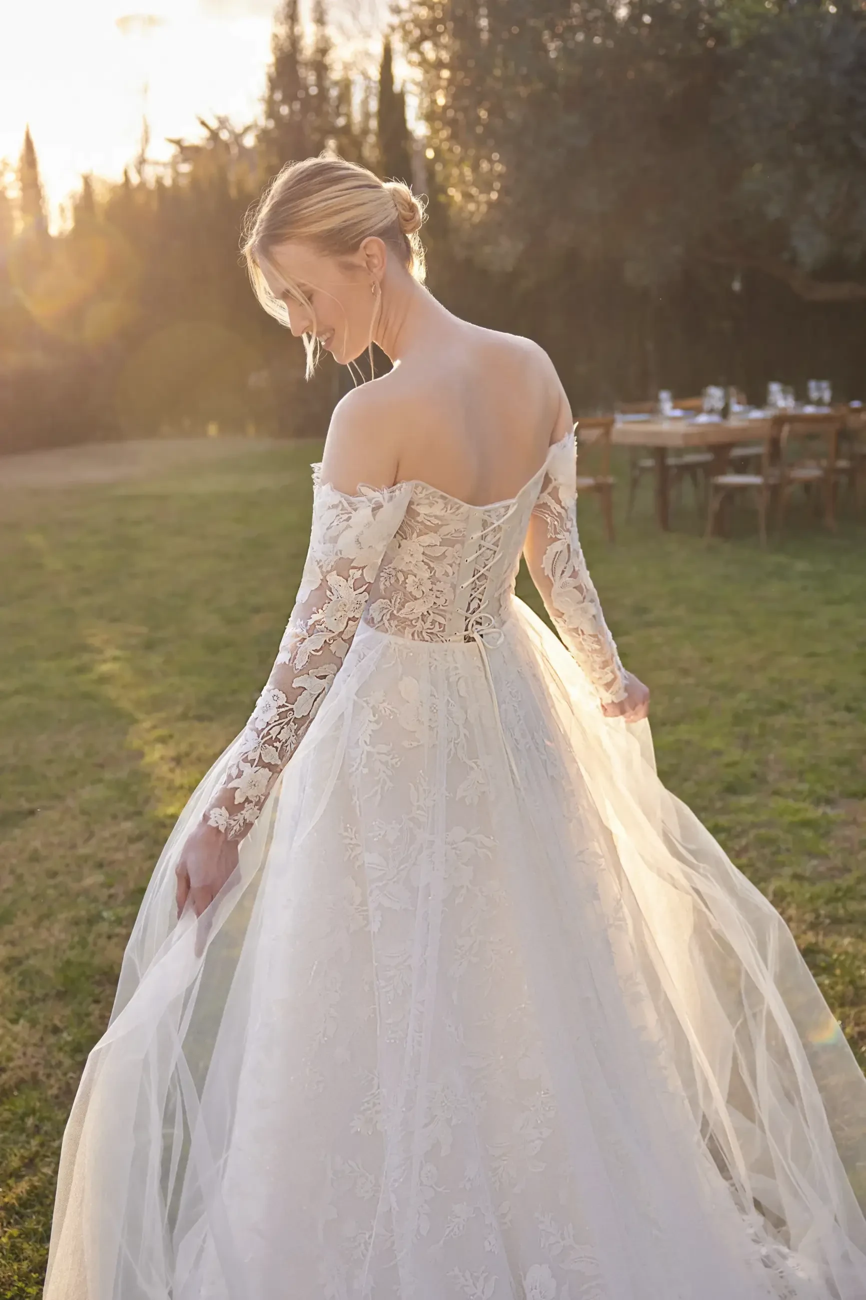 A bride in a beautiful lace wedding dress stands outdoors, looking back over her shoulder as the sun sets.