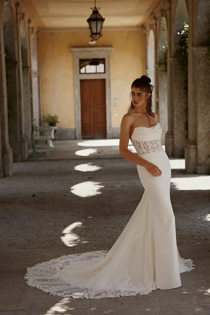 A woman wearing a fitted white wedding dress, standing in a hallway with arches and soft lighting.