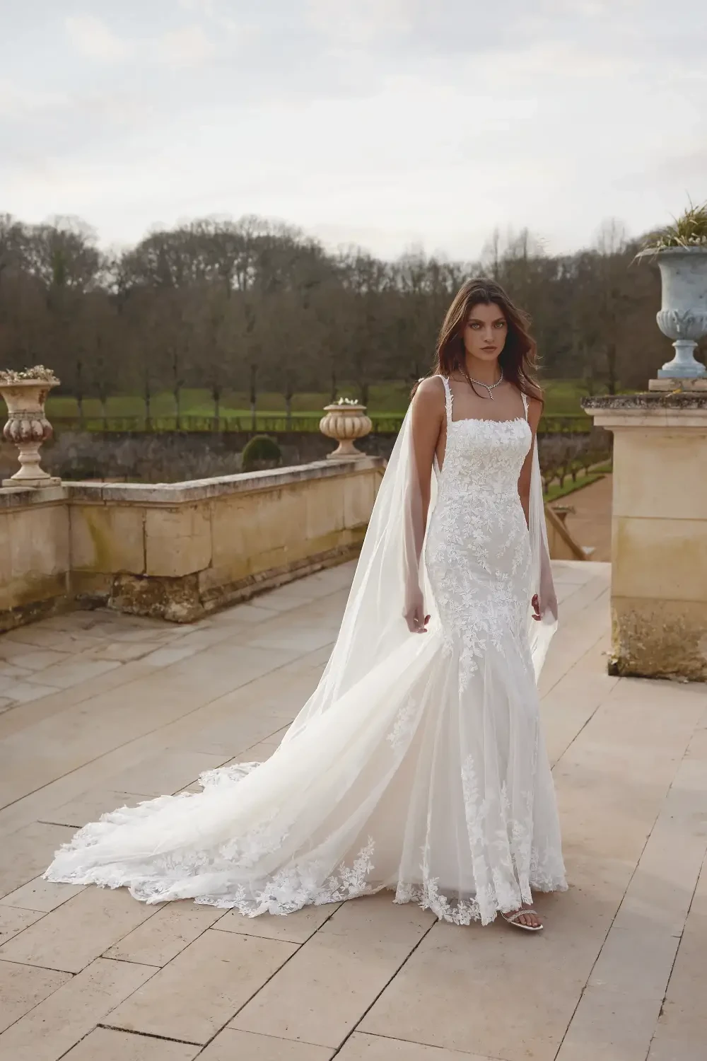 A bride wearing a beautiful lace wedding dress with a long train and a veil, standing outdoors on a pathway with trees in the background.