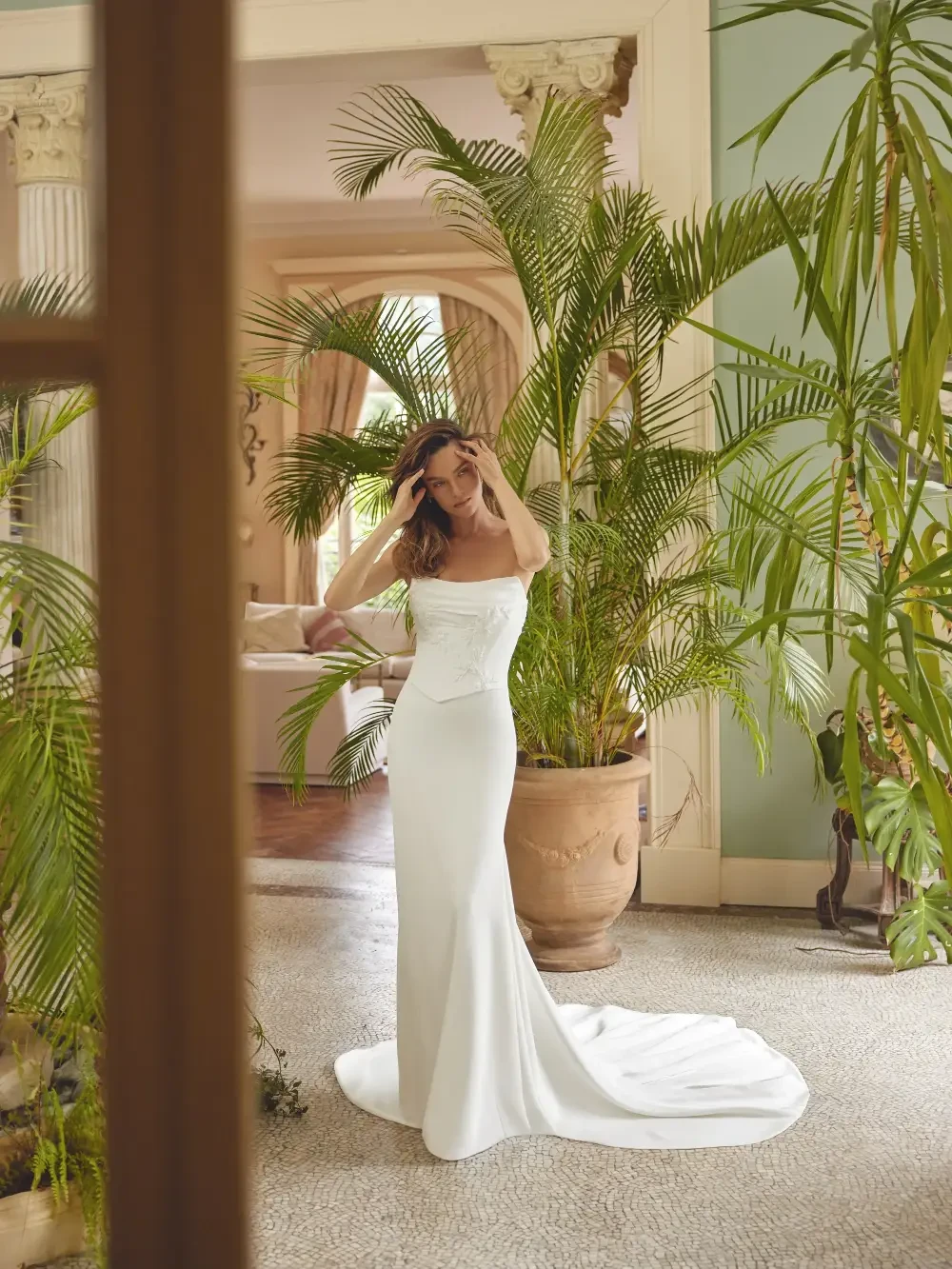 A woman in a white off-the-shoulder dress stands amidst greenery in an elegantly decorated room.