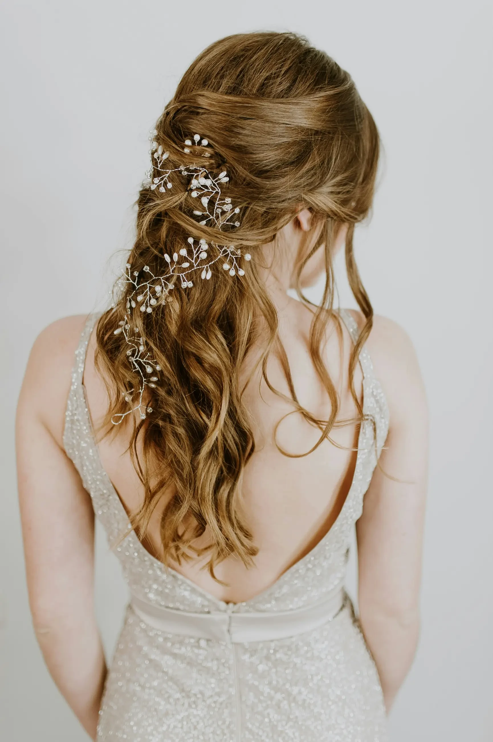 A woman with long, wavy hair styled with floral hair accessories, wearing a sleeveless silver dress. The image captures her back and the details of her hairstyle.