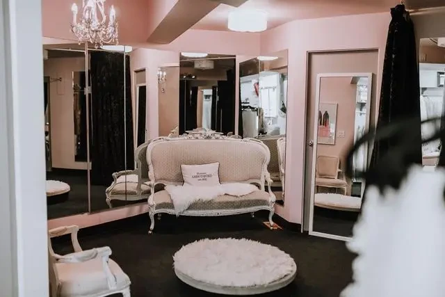 A stylish fashion fitting room with pink walls, a chandelier, mirrors, and elegant furniture including a vintage-style couch and round rug.