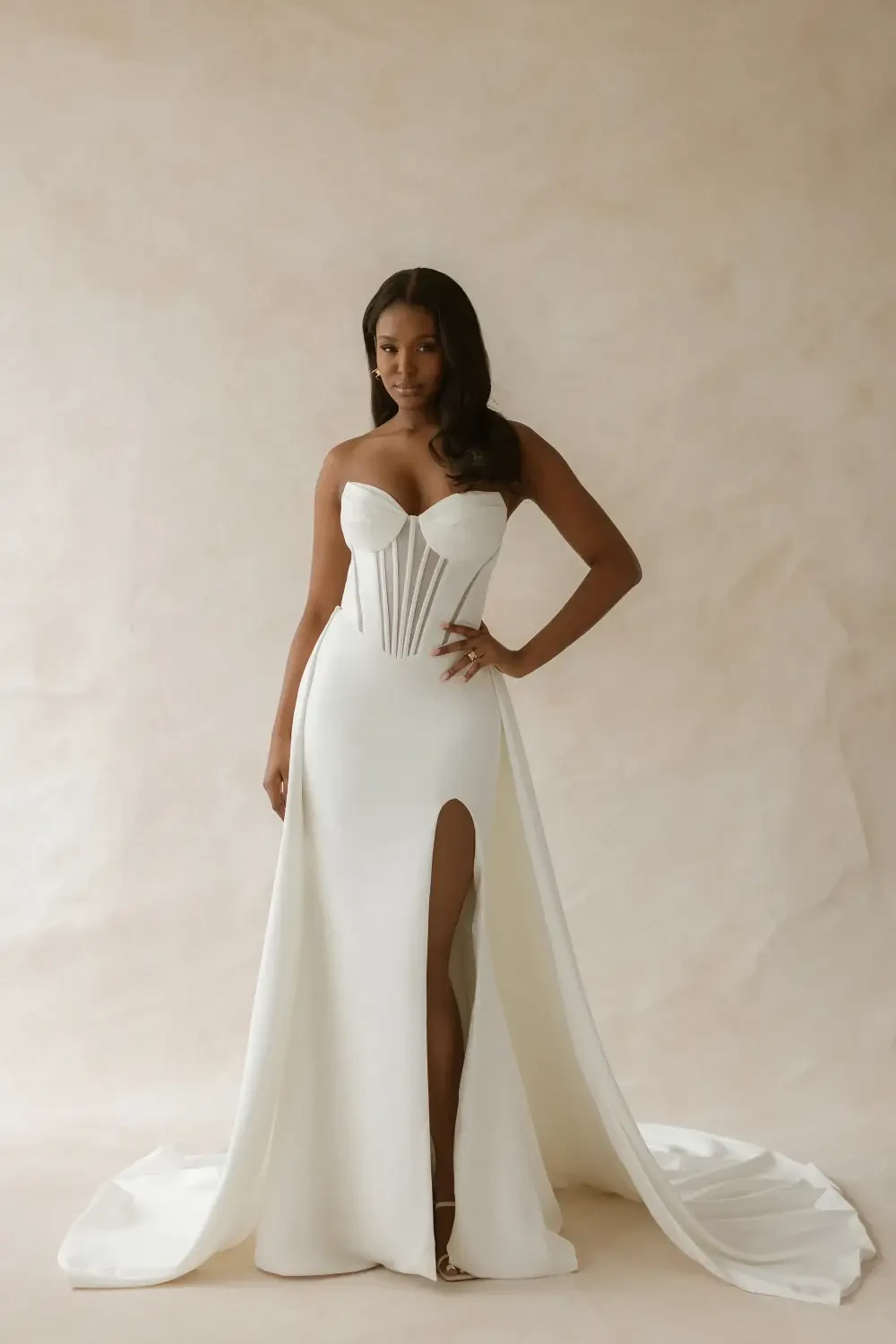 A woman in a strapless white gown with a high slit and a flowing train, posing confidently against a neutral background.