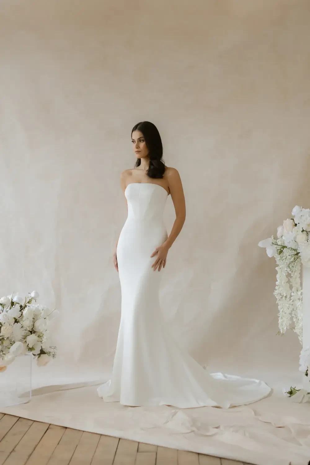 A woman in a sleek, strapless white gown standing against a neutral backdrop, with floral arrangements nearby.