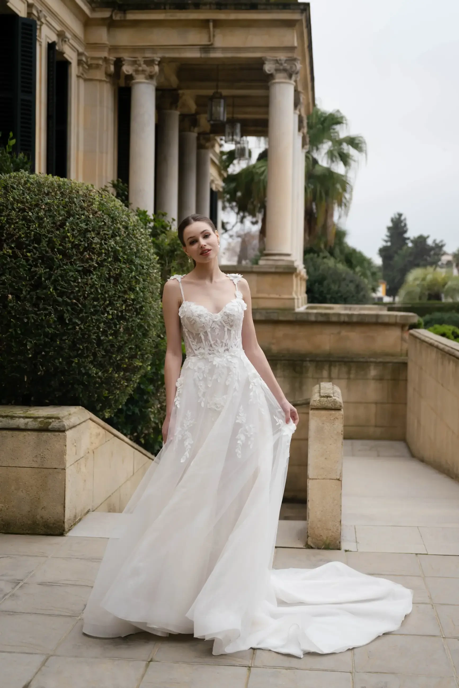 A woman in a white, lace wedding dress stands outside a classical building with columns. She looks serene, surrounded by greenery on a stone path.