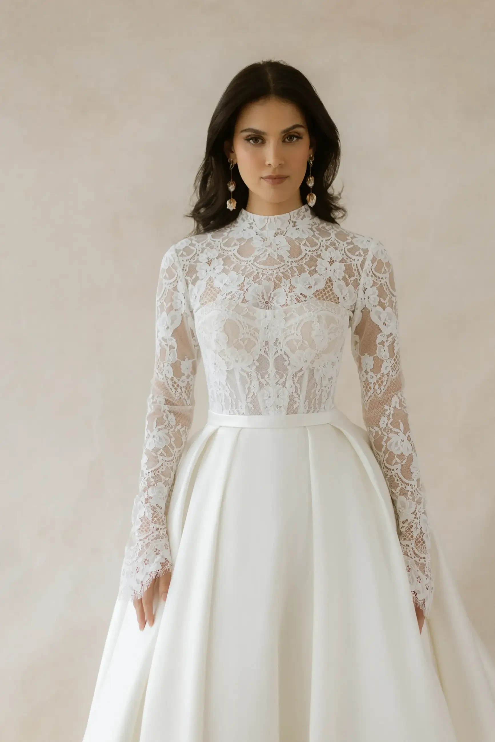 A woman in an elegant white wedding dress with lace sleeves and a high neckline. She wears statement earrings and has a calm, poised expression.