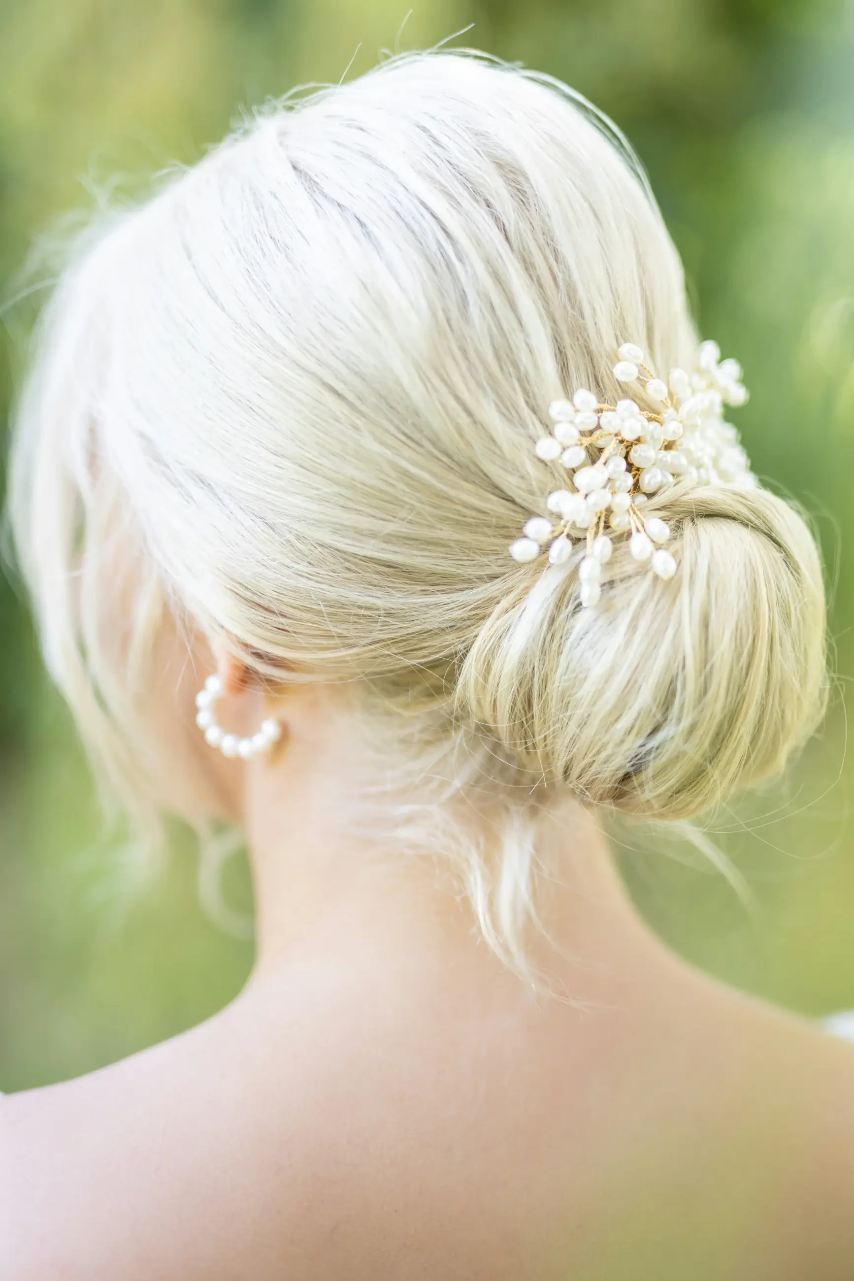 A close-up of a woman's hairstyle, showing a low bun adorned with hair accessories and pearl earrings. The perspective is from behind, focusing on the elegant hairdo and decoration.