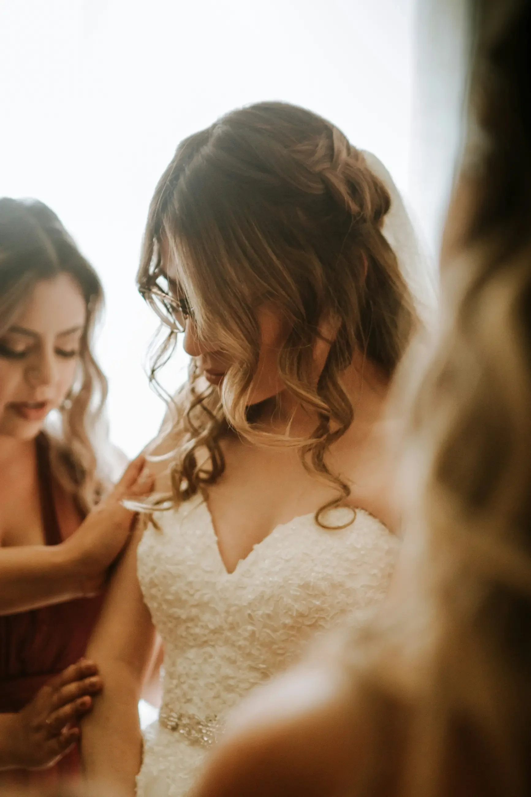 A bride in a wedding dress is adjusting her attire while surrounded by women are helping her prepare for the ceremony.