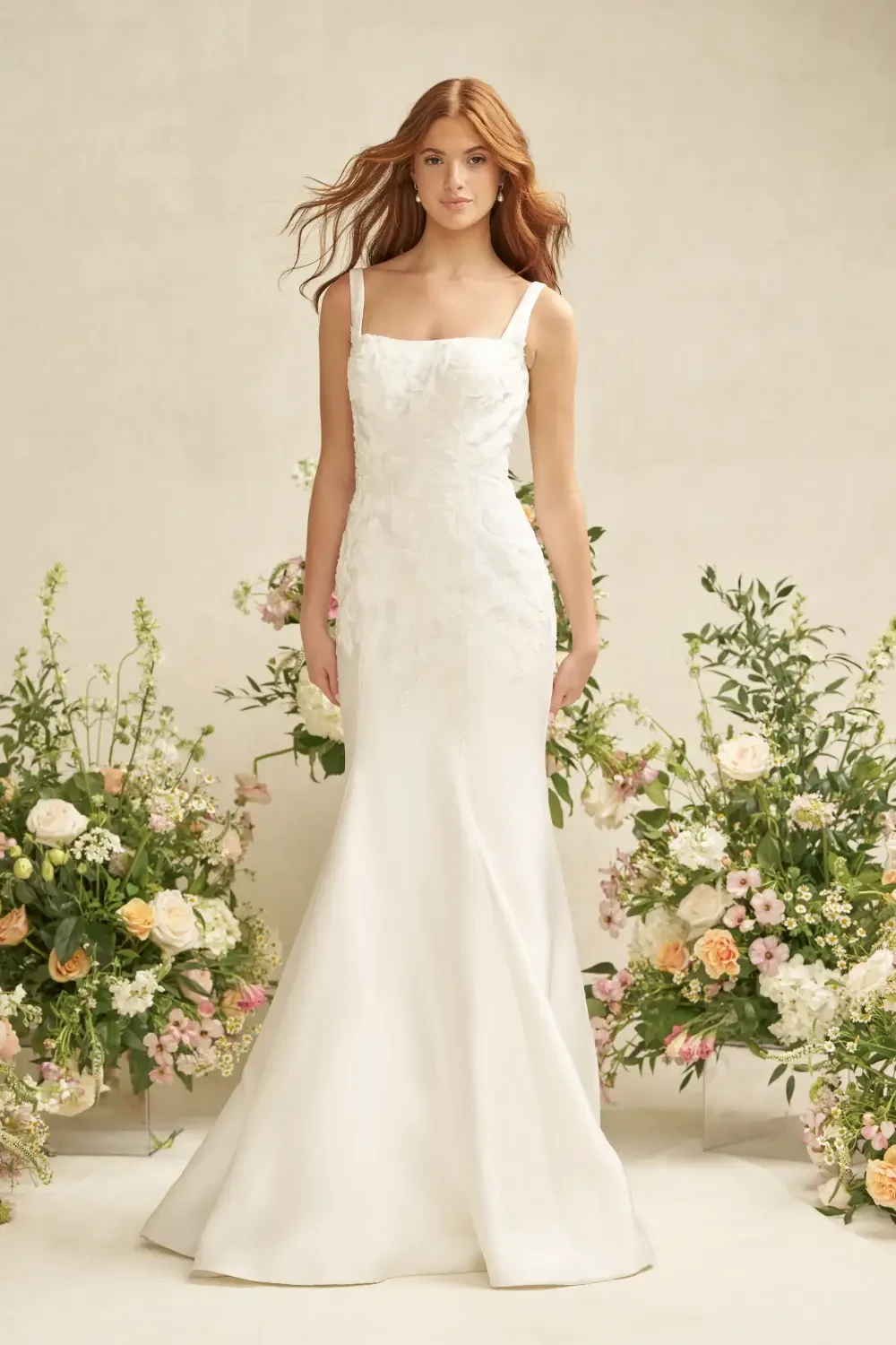 A model wearing a fitted white wedding dress with floral details, standing in front of a backdrop adorned with colorful flowers.